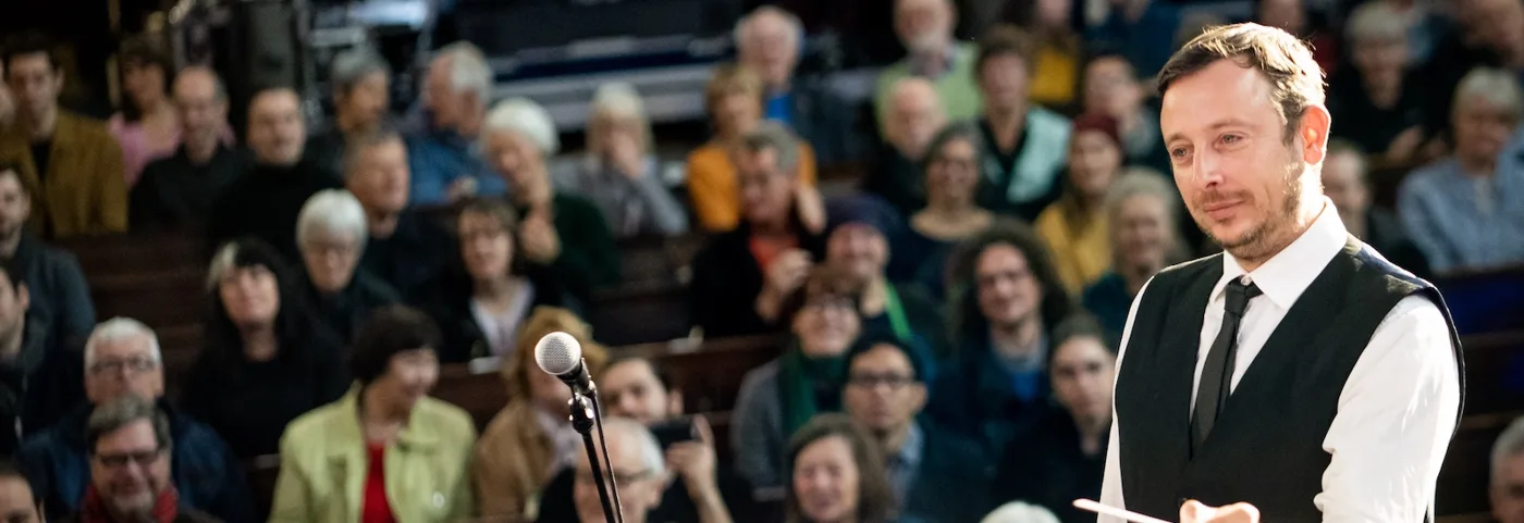 Ian Watson, conductor of London Accordion Orchestra, facing the orchestra with his baton raised, an audience seated behind him